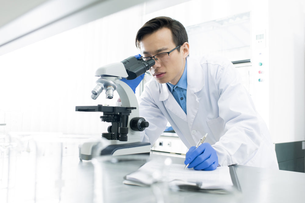 A male scientist in a white lab coat and blue gloves looks into a microscope while holding a pen and taking notes in a laboratory.