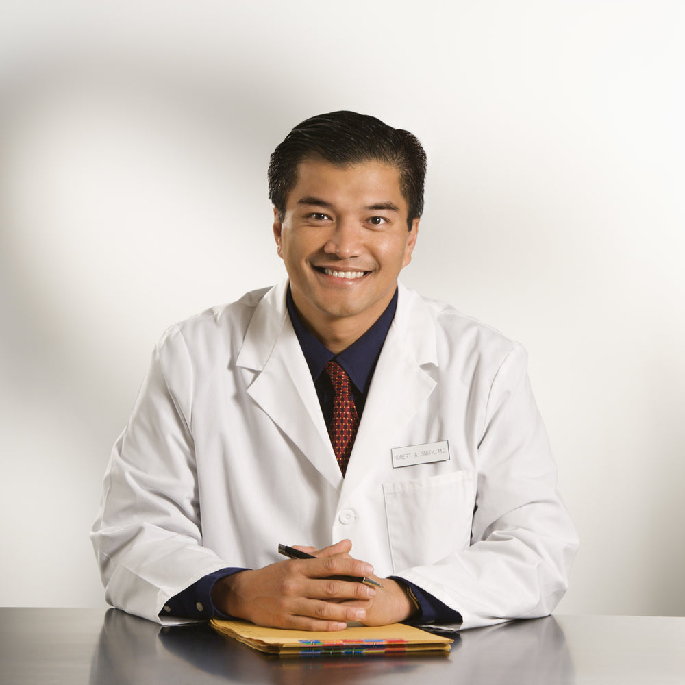 A smiling male doctor in a white lab coat sits at a desk with a folder, holding a pen.